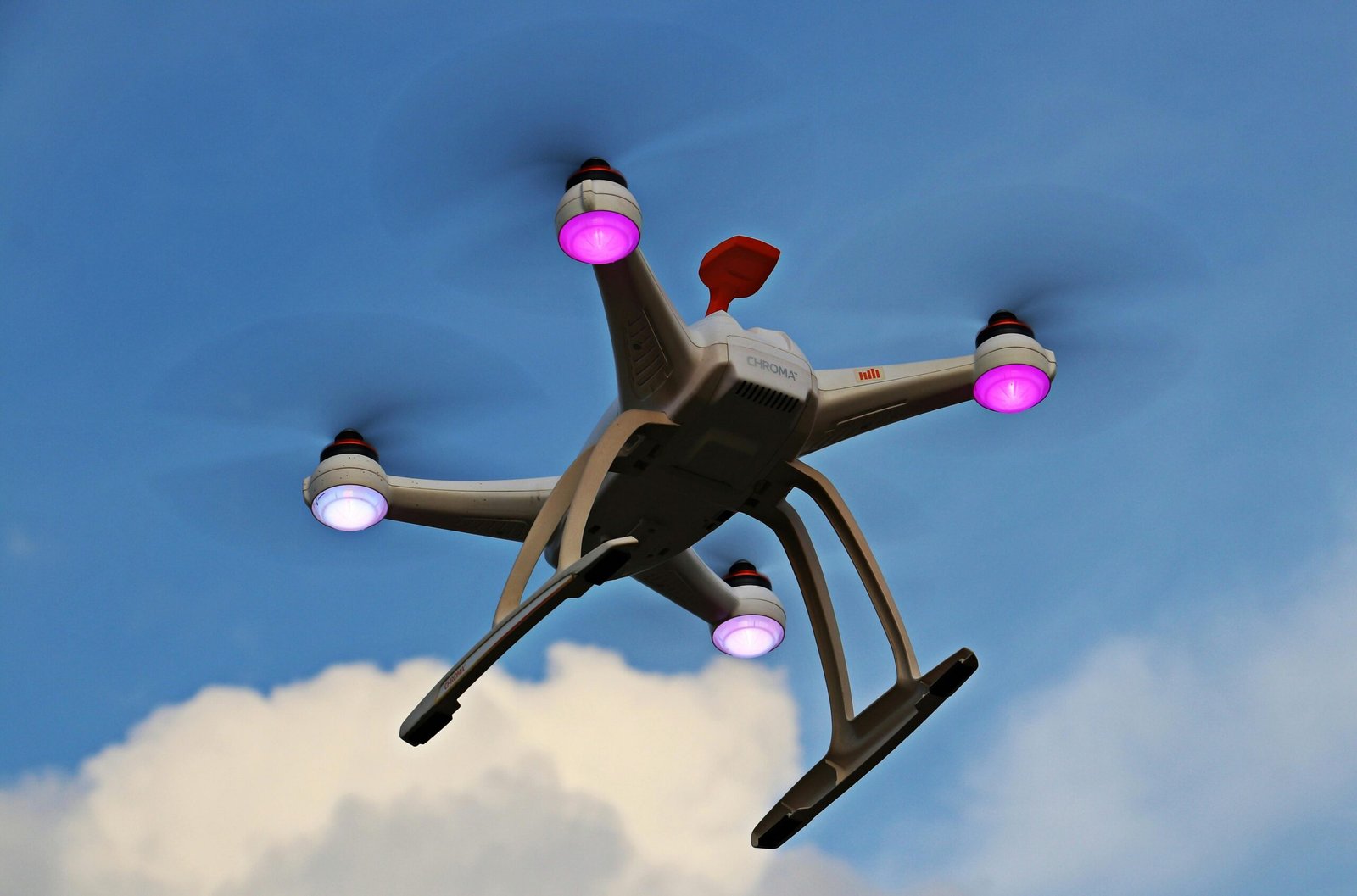 A quadcopter drone with propellers and lights flying high against a blue sky with clouds.